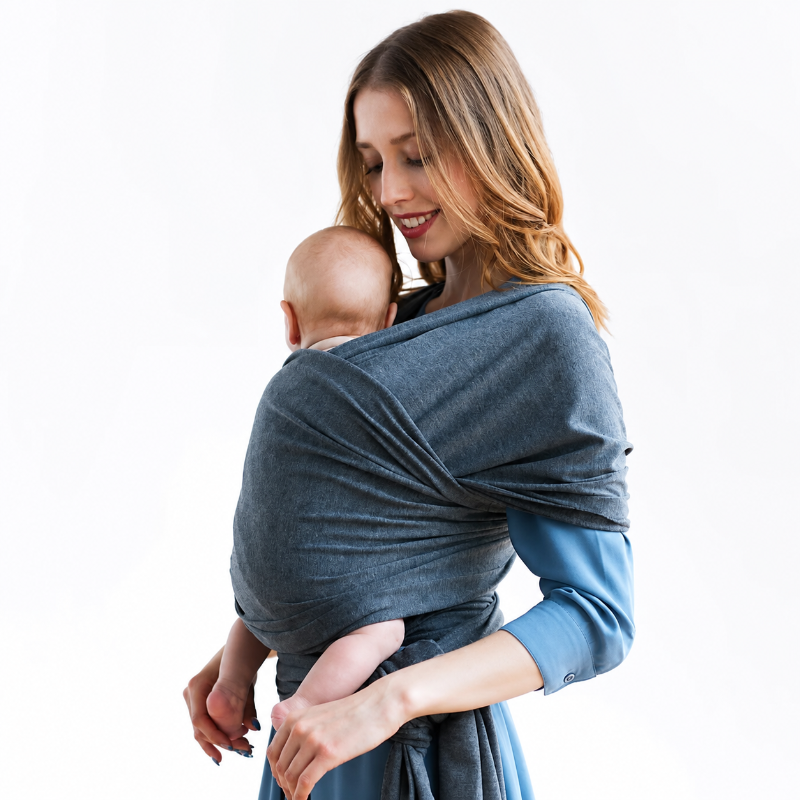 A woman wearing a dark slate blue wrap carrier with a baby on her back, shown against a plain white background.